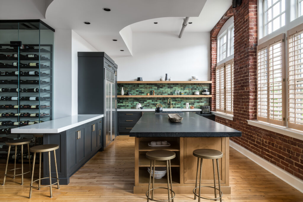 Luxury modern kitchen remodel featuring dark custom cabinetry, large island, green tile backsplash, exposed brick, and natural light filled windows.